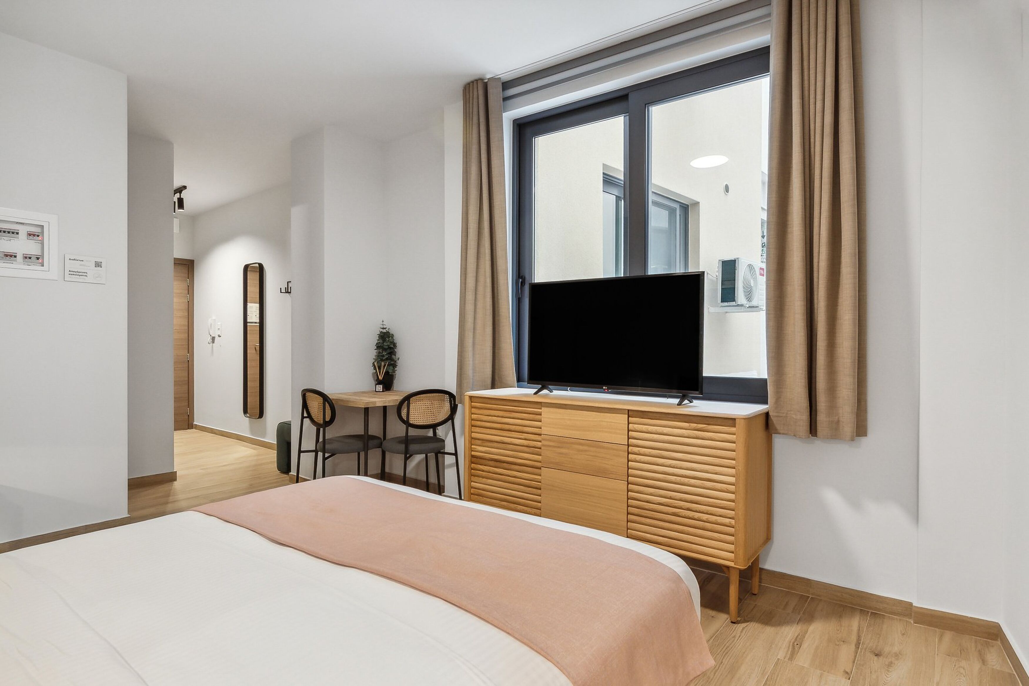 A modern hotel room with a large bed, a TV on a wooden dresser, a small table with chairs, and a window with curtains.