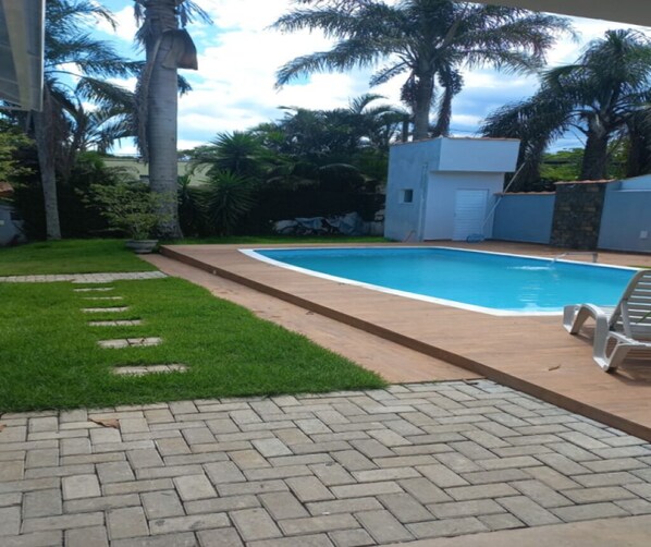 A swimming pool with a paved deck, surrounded by palm trees and a small building.
