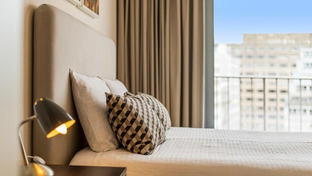 Una habitació d'hotel amb un llit, una tauleta de nit amb una làmpada i vista a la ciutat a través de grans finestres.
