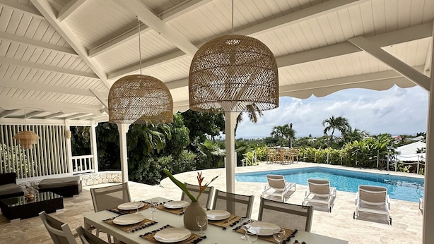 Uma área de refeições ao ar livre com vista para a piscina, mobiliário branco e luminárias trançadas.