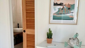 Um banheiro com porta de madeira, pia, cacto em vaso e um quadro na parede.