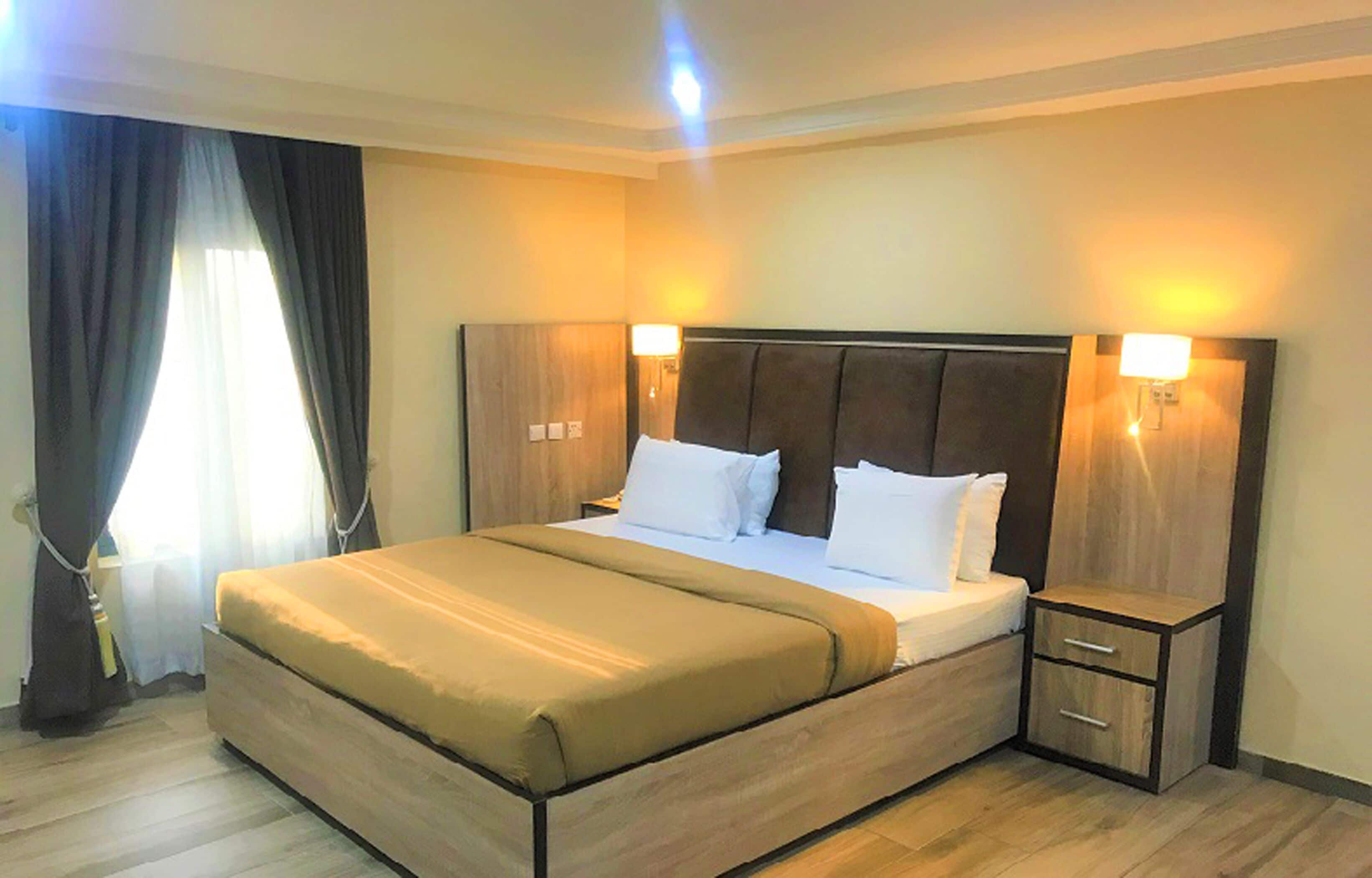A modern hotel room with a large bed, wooden headboard, bedside tables, and a nightstand.