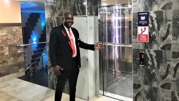 A man in a black suit with a red tie standing next to an elevator with a digital display showing the number 3. - CASONI INTERNATIONAL HOTELS AND SUITES (Port Harcourt)
