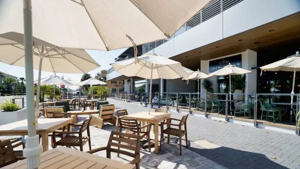 An outdoor cafe area with wooden tables and chairs, large umbrellas, and a modern building in the background. - Ocean Seaside Escape B
 (Jacksonville Beach)
