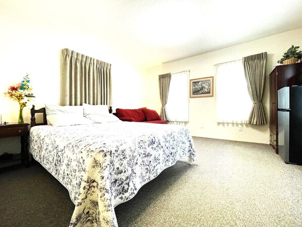 A hotel room with a bed, a nightstand, a lamp, a dresser, and a refrigerator. - Apt. 3 - Ensuite Bath, Roku, Fridge/Micro, Queen (Paulding)