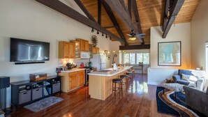 A spacious kitchen with wooden beams, a large island with stools, a flat-screen TV, and a dining area with a sofa and rug. - Dollar Hill II # 25 by Hauserman Rental Group (Tahoe City)