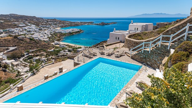 Zona de piscina con una piscina azul cristalina, escalones de piedra y vistas al mar y edificios al fondo.