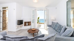 Un salón moderno con un sofá gris, una mesa de centro redonda de madera y un televisor en una pared blanca. Se aprecia una pared de piedra y vista al mar a través de una puerta abierta.