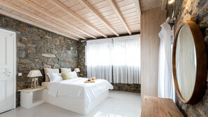 Une chambre avec un mur en pierre, des poutres en bois au plafond, un lit avec du linge de maison blanc, une table de chevet avec une lampe et un grand miroir.