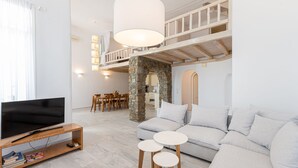 A modern living room with a TV, sofa, and two small tables. There is a staircase leading to an upper level with a balcony. - Luxury VillaThelgoMykonos IV, amazing sea View! (Mykonos)