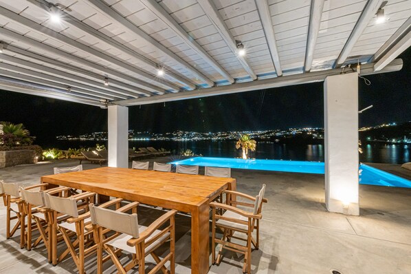 Une terrasse extérieure avec une table et des chaises en bois, une piscine et une vue sur le paysage urbain la nuit.