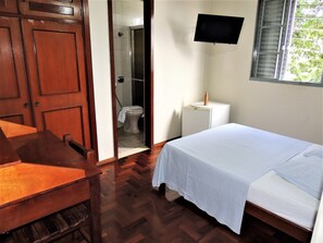 A room with a wooden desk, a bed with a white bedspread, a small white cabinet, a television mounted on the wall, and a door leading to a bathroom.