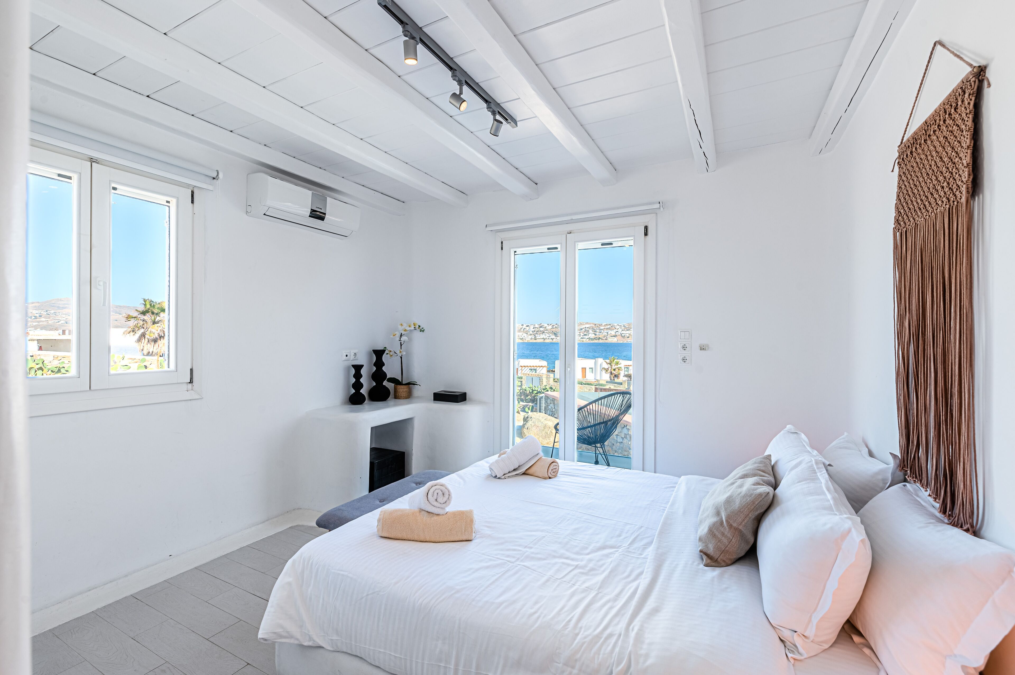 Ein heller, weißer Raum mit einem Bett, einem kleinen Tisch und einem Stuhl. Zwei Fenster bieten Blick auf das Meer.
