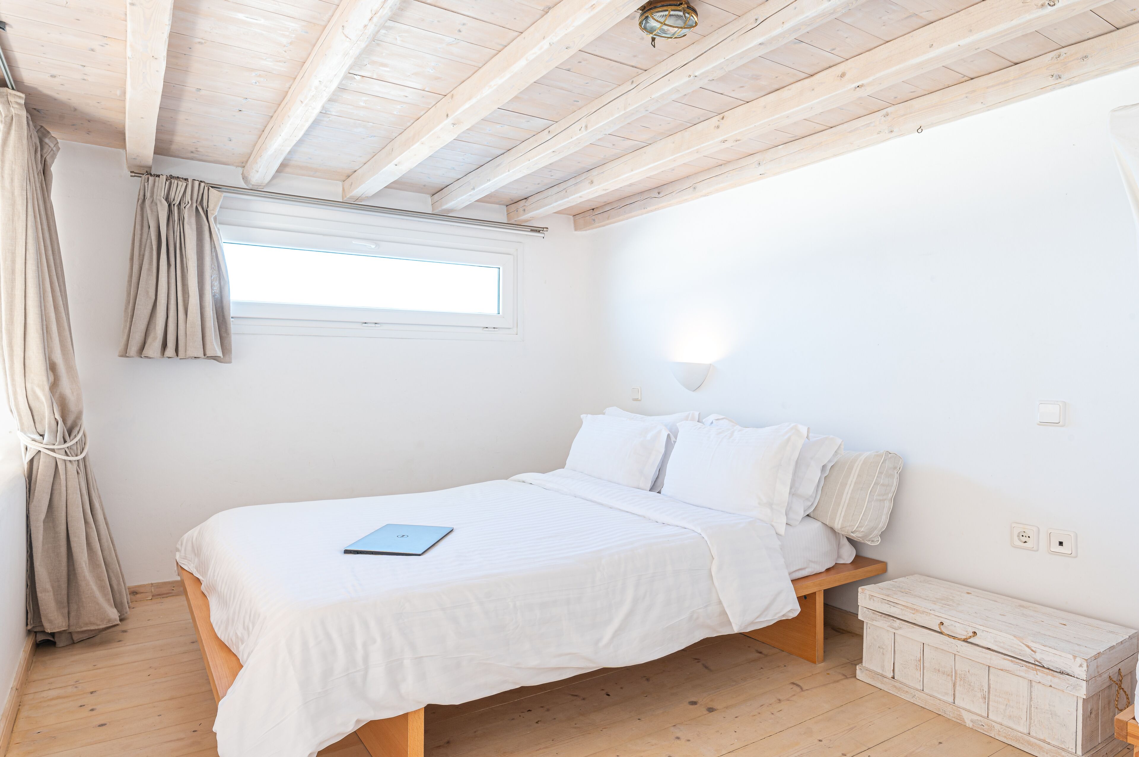A bedroom with a wooden bed, a bedside table, a window with curtains, and a light fixture on the wall.