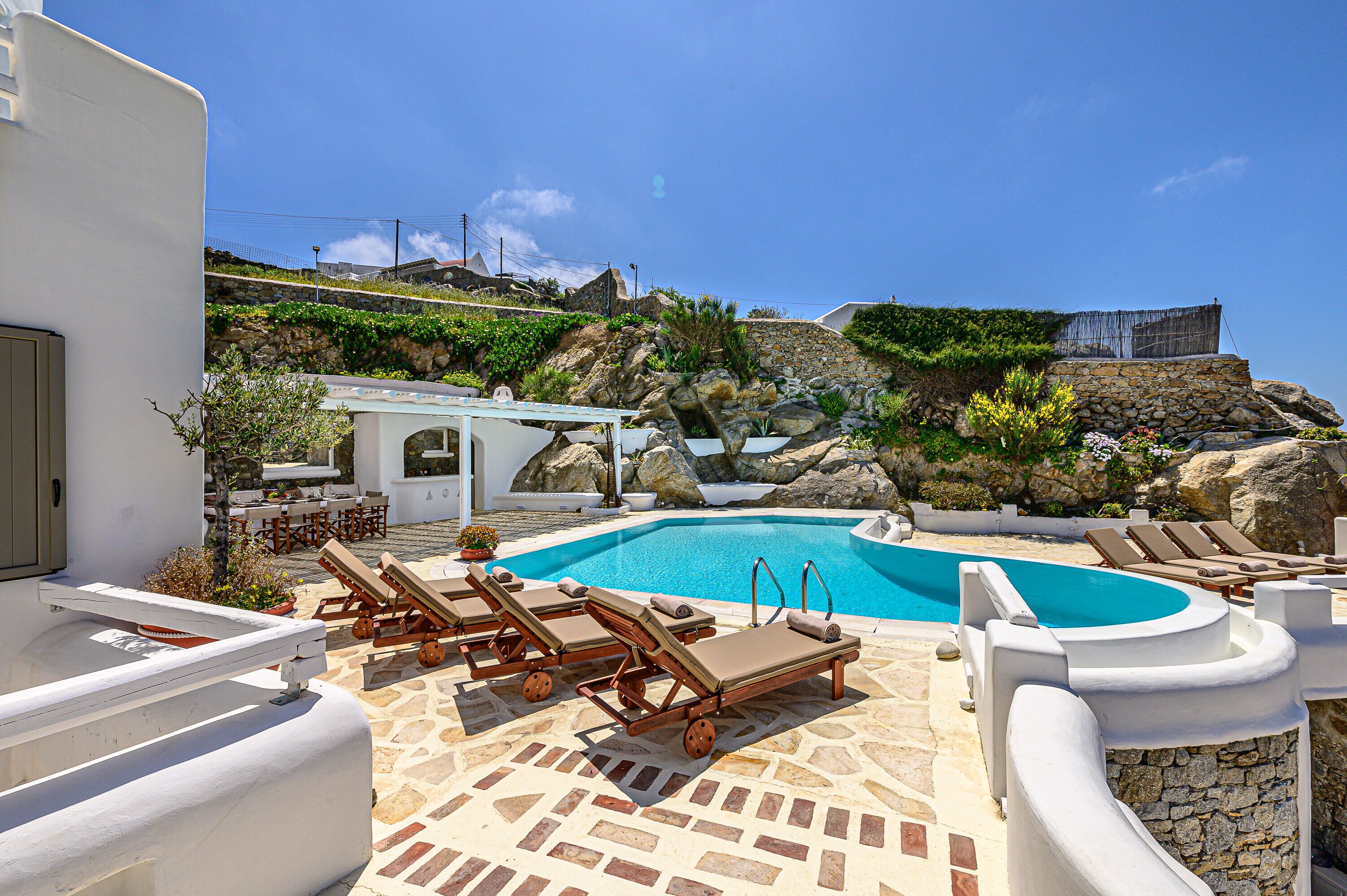 A pool area with lounge chairs, a stone-tiled deck, and a view of a hillside with a building in the background.