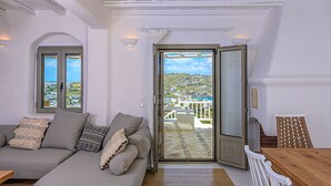 A modern living room with a sofa, a wooden table, and a view of a balcony. - 9 Muses Villa Thalia 5 BR with private pool! (Mykonos)