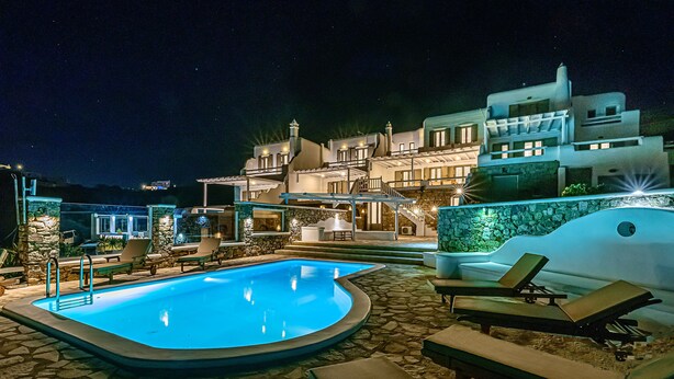 A nighttime swimming pool area with lounge chairs, a stone-tiled deck, and a modern building with multiple balconies and lights. - 9 Muses Villa Thalia 5 BR with private pool! (Mykonos)