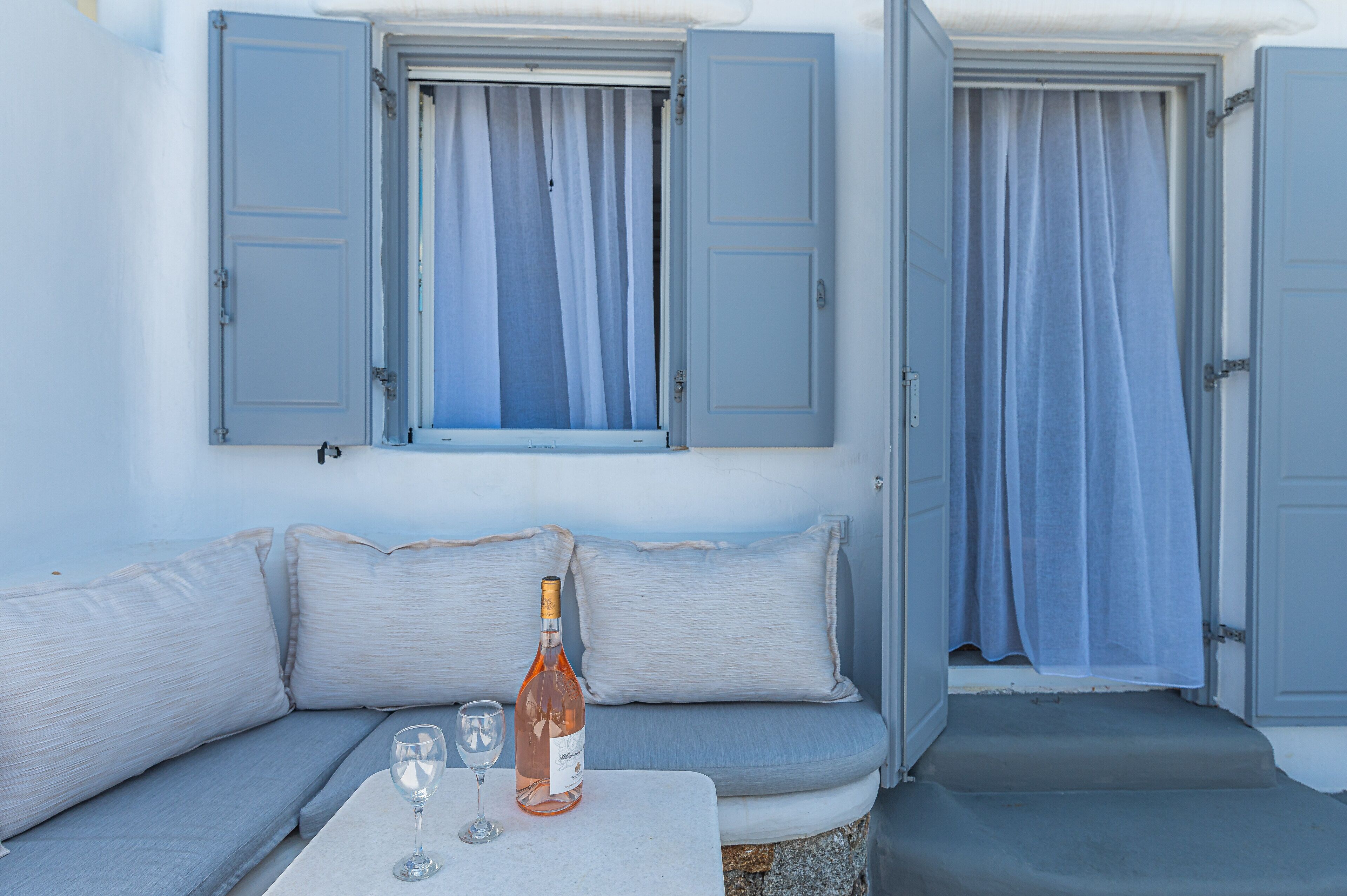 A room with a light blue sofa, a table with a bottle and glasses, and two windows with blue shutters.