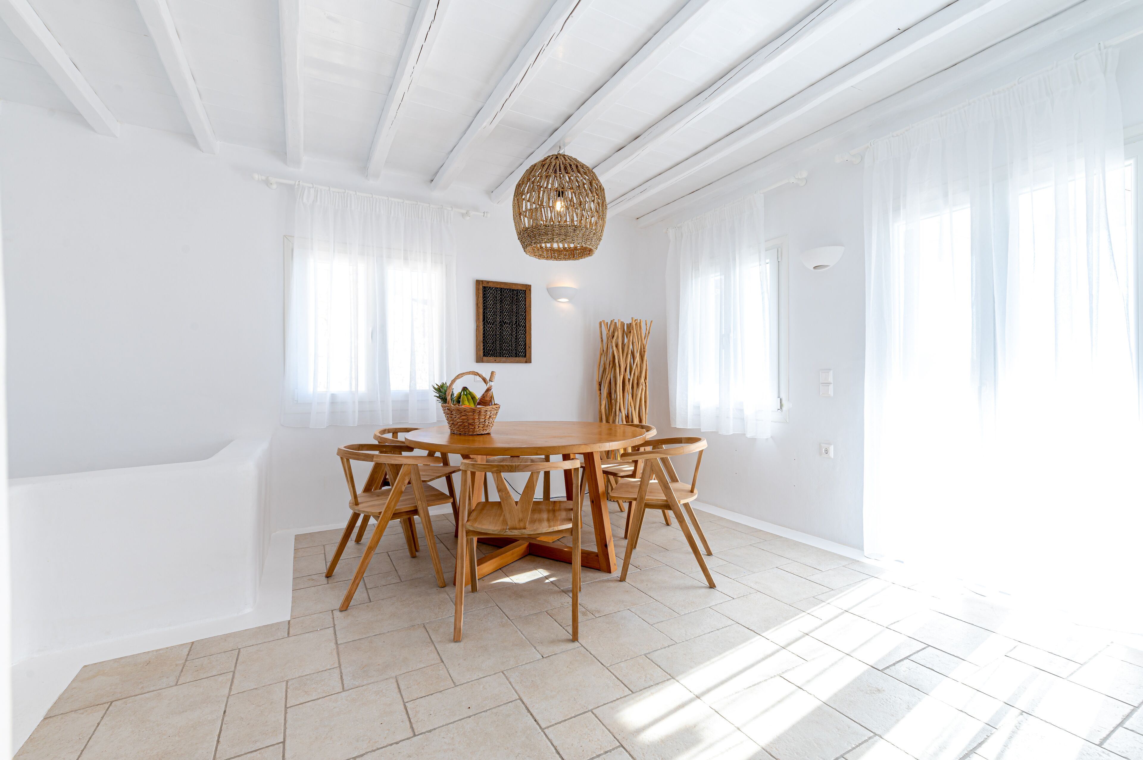 A bright dining area with a round wooden table and chairs, a woven light fixture, and large windows with sheer curtains.