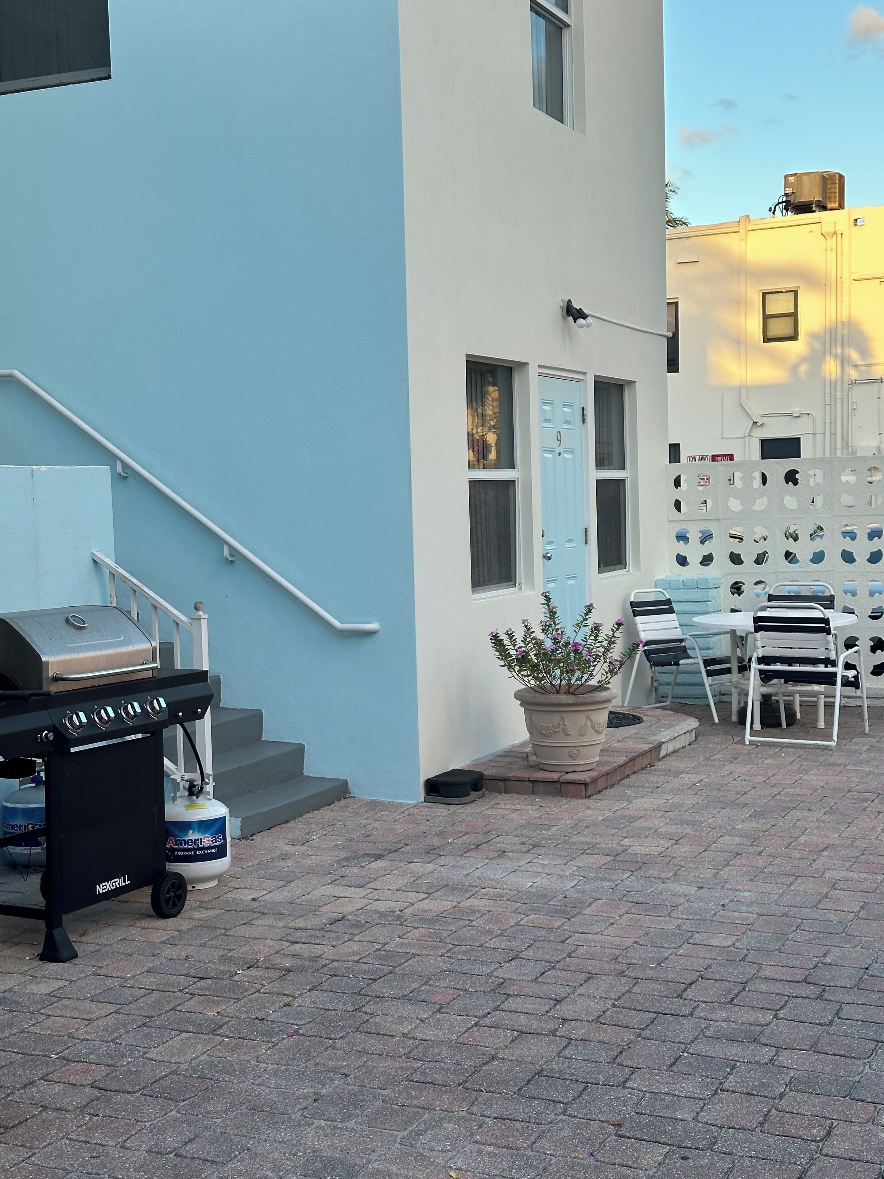 A residential building with a blue exterior, a front door, a staircase, a barbecue grill, and outdoor seating with chairs and a table.