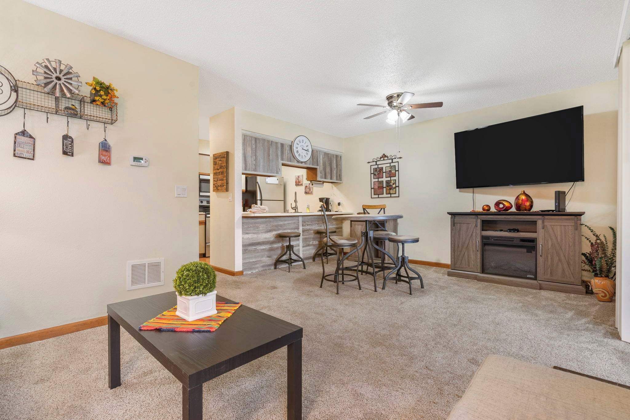 A modern living room with a dark wooden coffee table, a kitchen area with a bar, and a flat-screen TV mounted on the wall.