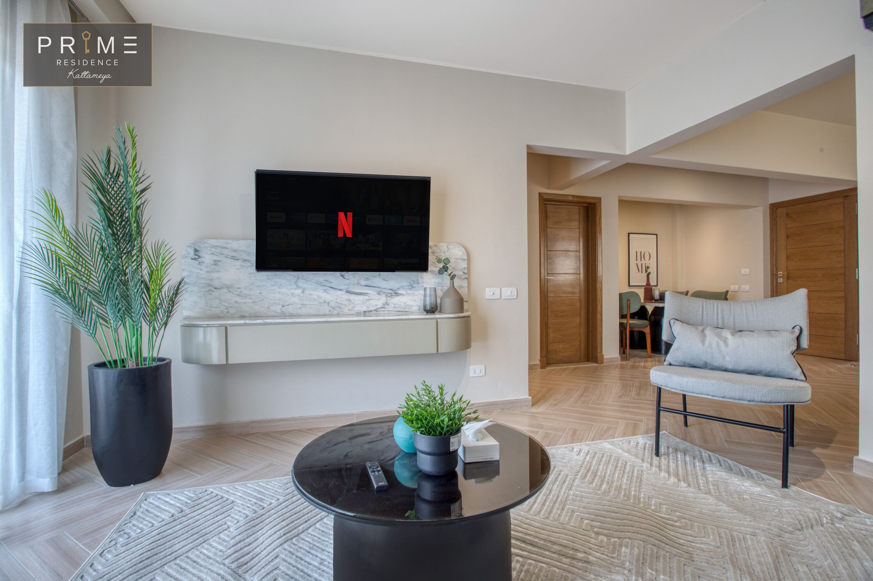 Un salon moderne avec un téléviseur à écran plat fixé à un mur blanc, une table basse noire agrémentée d’une plante, et un canapé gris avec un coussin.
