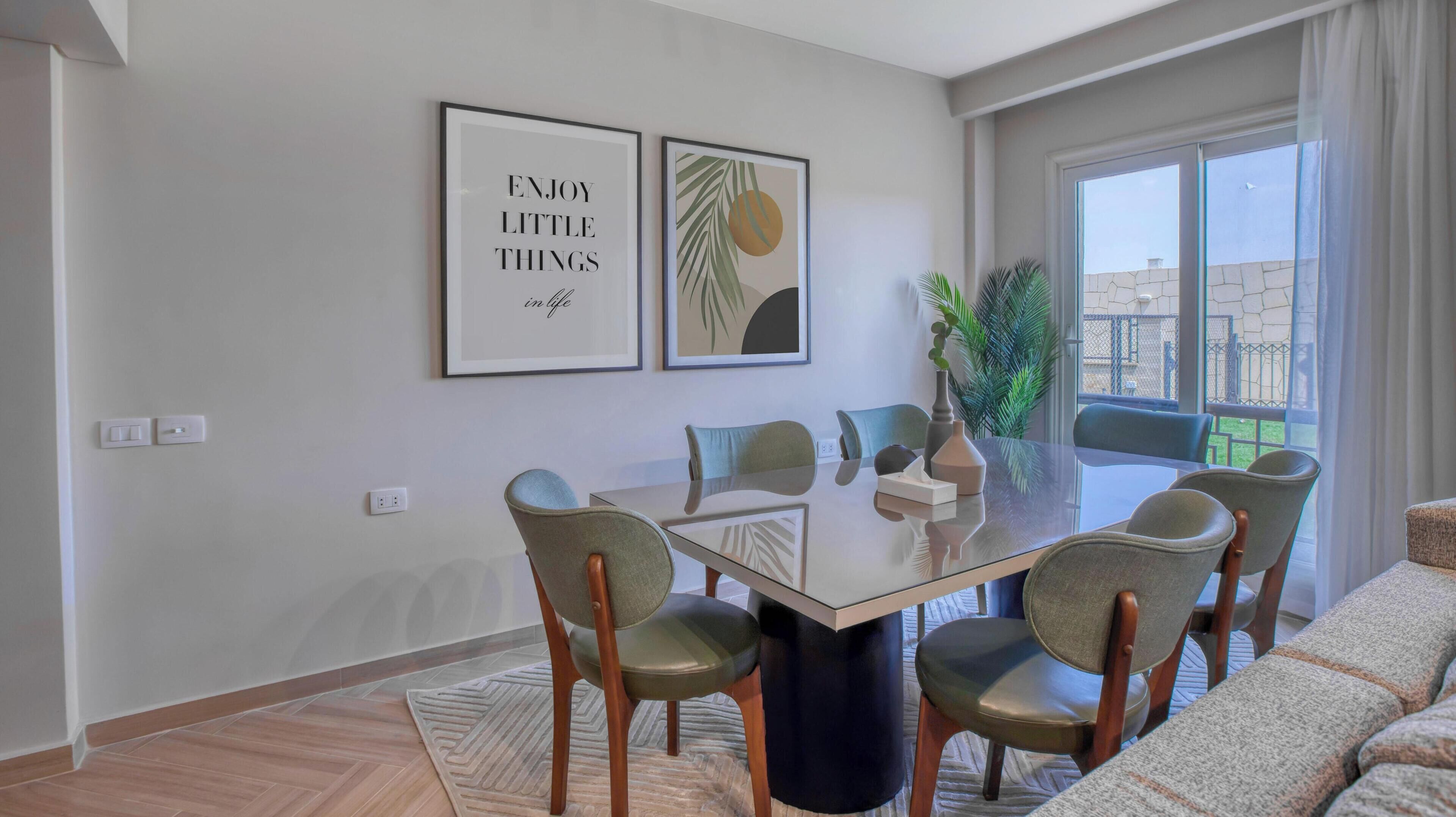 A dining area with a glass table, six chairs, two framed artworks, and a potted plant.