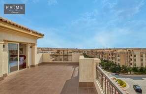 Ein Wohngebäude mit Fliesenbalkon, ein Schlafzimmer mit Bett und Fenster sowie ein Blick auf andere Gebäude in der Ferne.