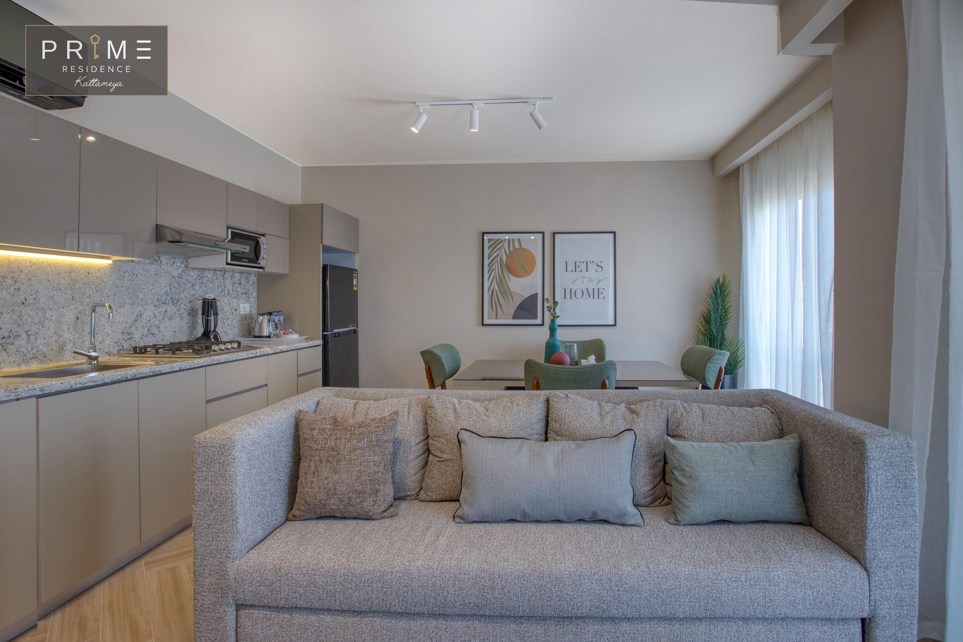 A modern living room with a grey sofa, a kitchen area with a built-in oven and microwave, and a dining area with a table and chairs.