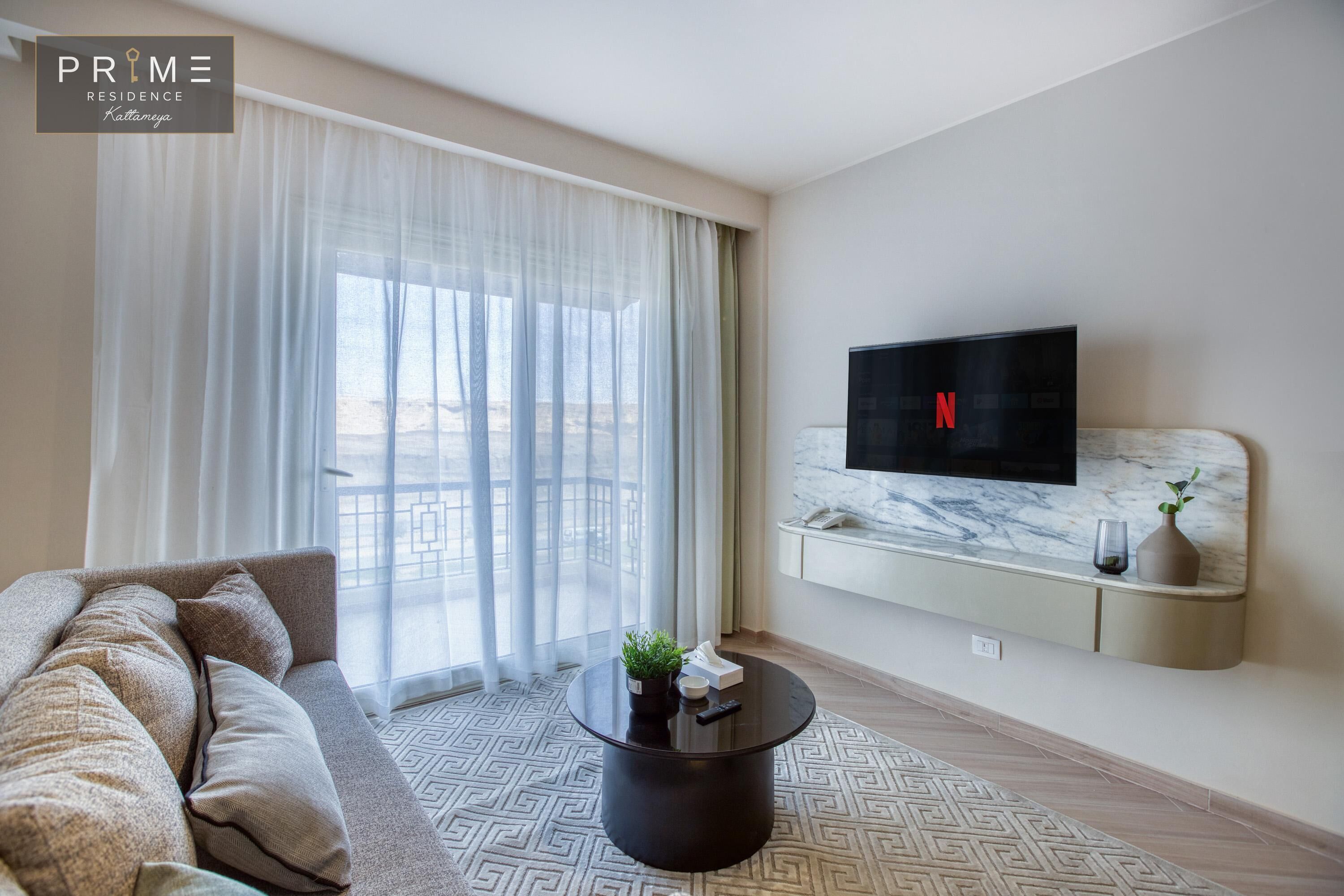 Una sala de estar moderna con un sofá, una mesa de centro, un televisor montado en una pared de mármol y un ventanal con cortinas translúcidas.