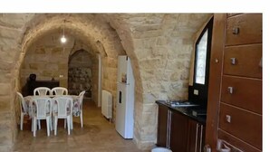 A stone-walled room with a dining table and chairs, a refrigerator, and wooden cabinets.