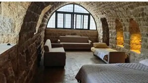 A cozy attic bedroom with a stone wall, a large arched window, a bed, a sofa, and a small table.