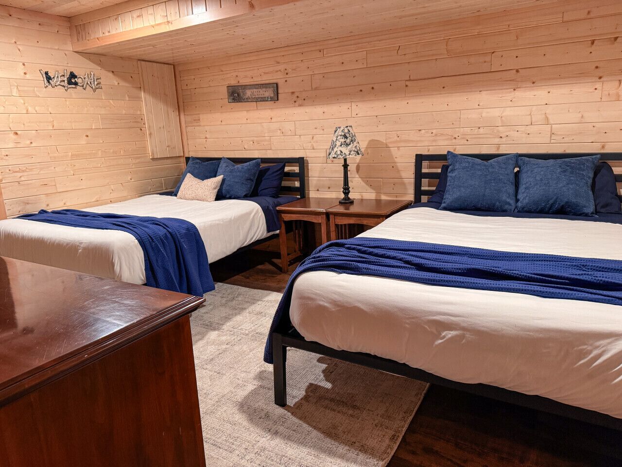 A room with two beds, wooden walls, a small table, and a lamp.