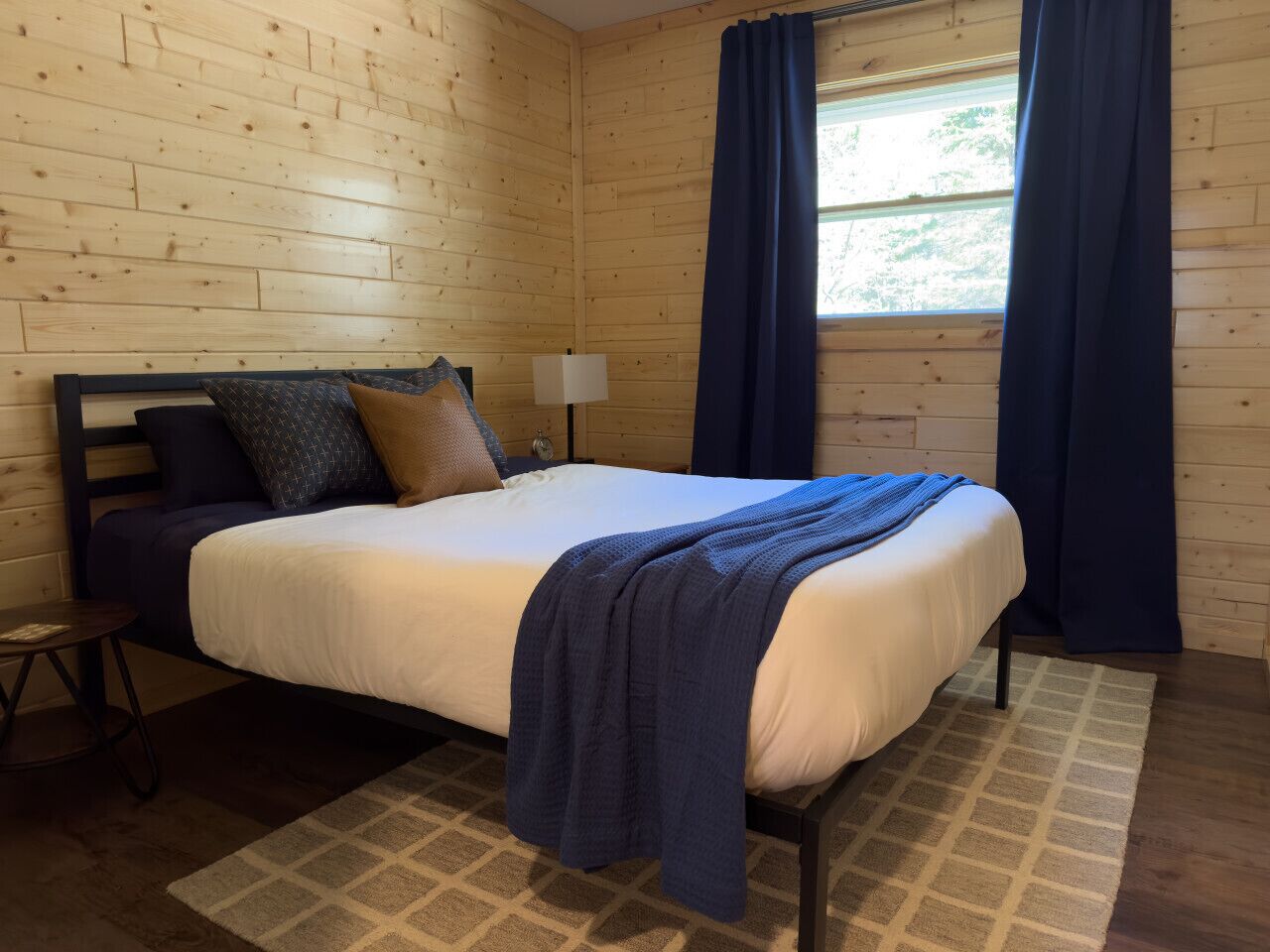 Ein Schlafzimmer mit einem Bett, einem Nachttisch, einer Lampe und einem Fenster mit Vorhängen.