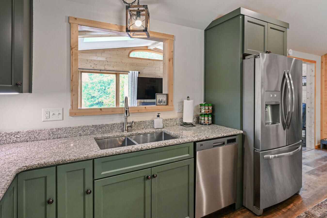 Una cocina con armarios verdes, un refrigerador de acero inoxidable, una encimera de granito, un lavabo y una ventana con vistas a la vegetación.