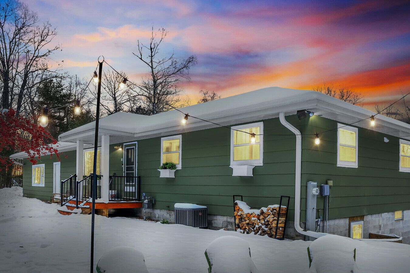 A green house with a porch, a window with a plant, and a stack of firewood.