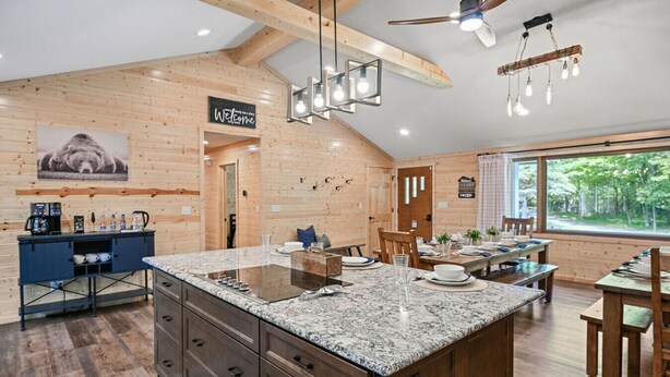 A modern kitchen with a large island, wooden walls, and a dining area with a view of the outdoors. - Elevated 6BR Lodge | On Trail | Speakeasy/Game Room (Eagle River)