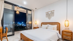 Una camera da letto con un letto grande, comodini in legno, una sedia in vimini e vista sulla città di notte attraverso una porta-finestra in vetro.