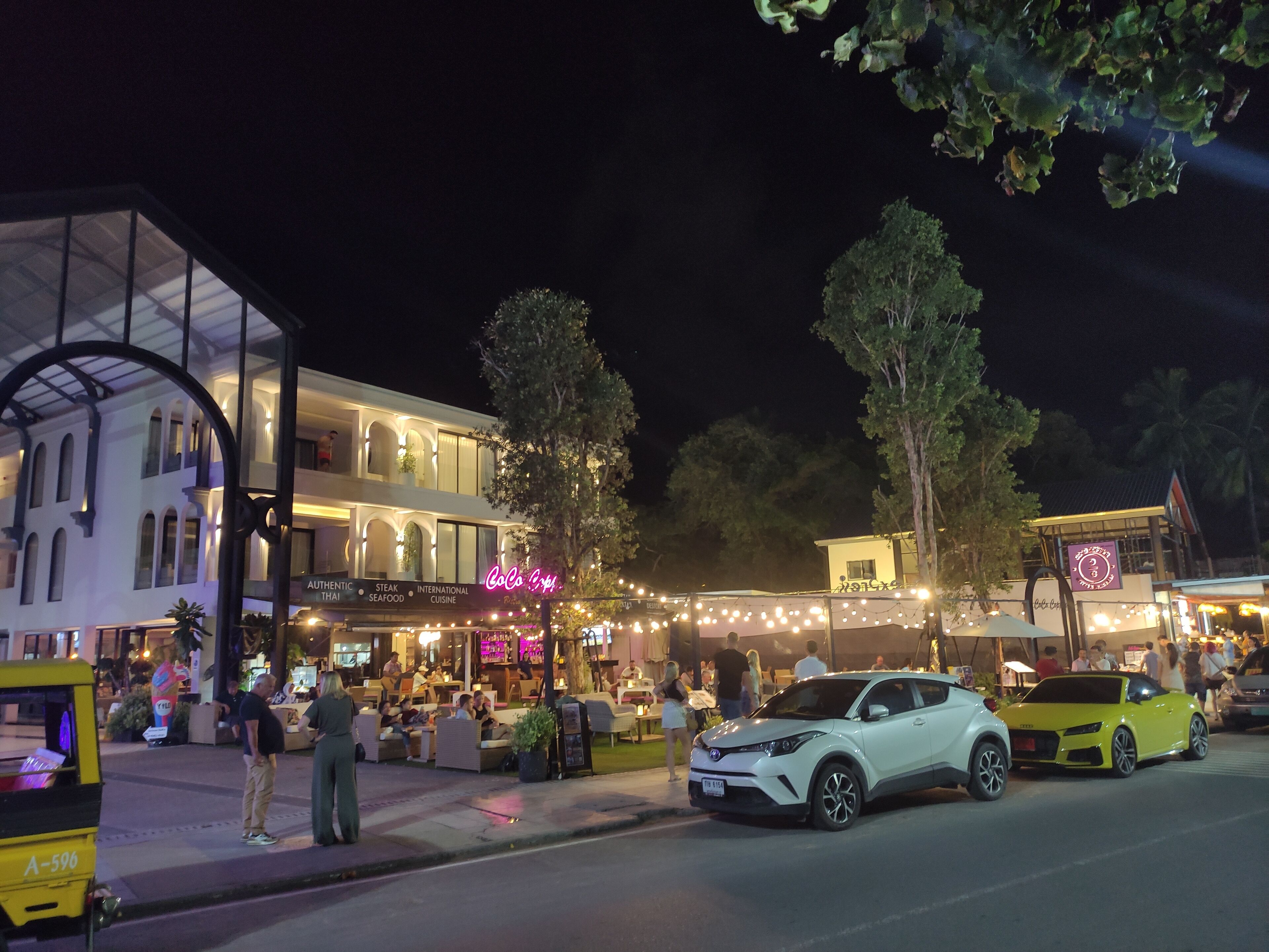 A street scene at night with a white car parked on the side of the road, a yellow auto-rickshaw, and people walking.