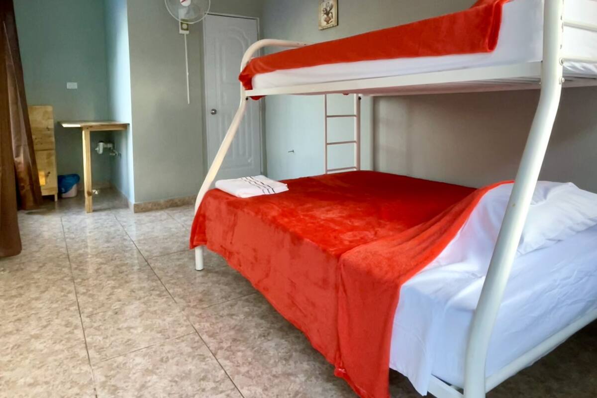 A bunk bed with a red bedspread, a white fan, and a small table with a chair in a room with tiled flooring.