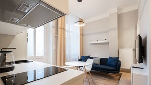 Una cocina moderna con una isla central, un comedor con mesa y sillas blancas, un sofá con cojines azules y un televisor sobre un mueble.