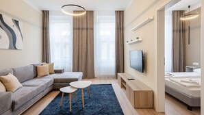 Una habitación de hotel moderna con un sofá gris, una mesita redonda pequeña, un televisor y una cama con cabecero de madera.