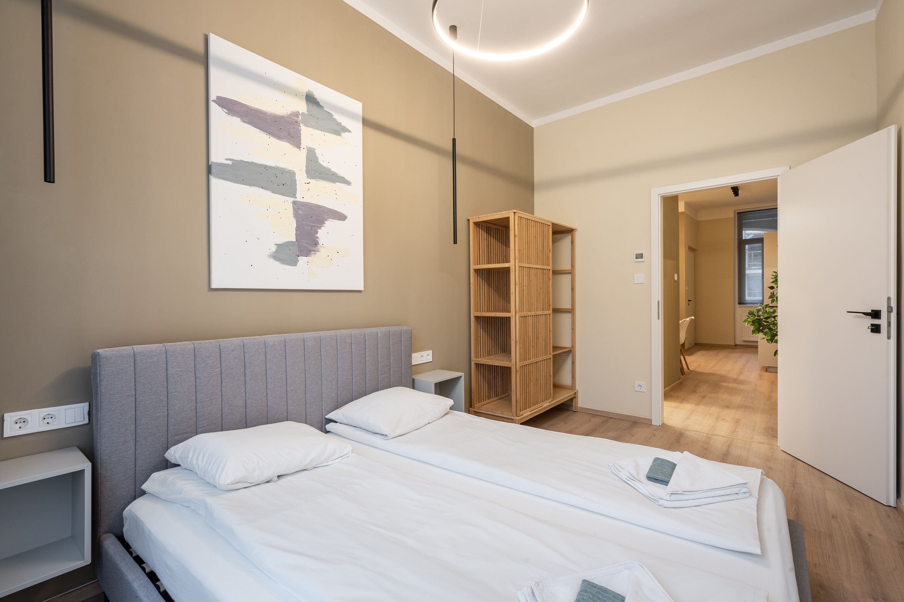A modern hotel room with a large bed, a bedside table, a wardrobe, and a view into the hallway.