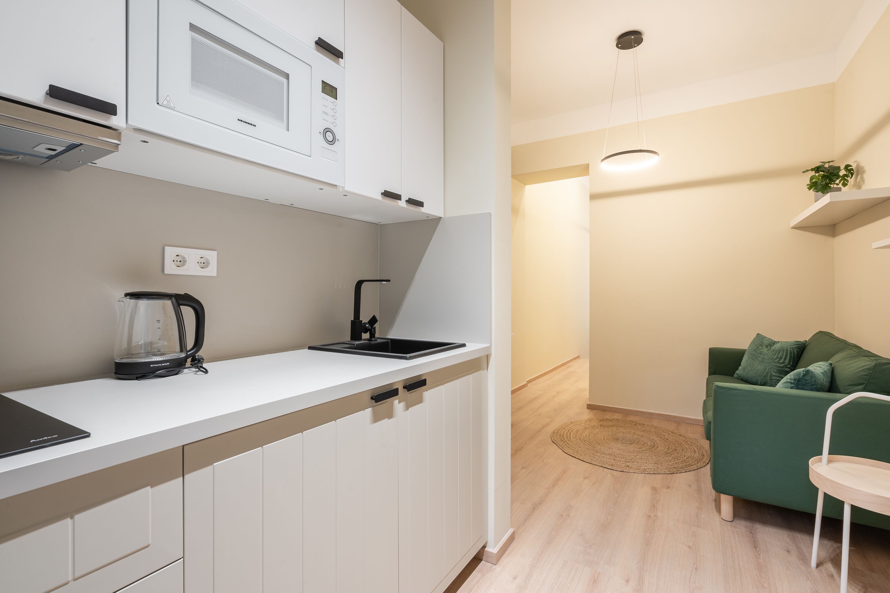A modern kitchen with white cabinets, a black kettle, and a green sofa in the adjacent living area.
