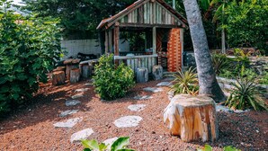 Eine rustikale Holzstruktur mit geneigtem Dach, umgeben von einem Garten mit verschiedenen Pflanzen und einem Kiesweg.