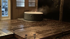 A hot tub on a wooden deck with a view of a tree and a building with windows. - Tiny House for two nestled under massive oaks in Southern Oklahoma. (Lone Grove)