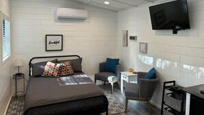 A bedroom with a bed, bedside table, chair, TV, and wall decorations. - Tiny House for two nestled under massive oaks in Southern Oklahoma. (Lone Grove)