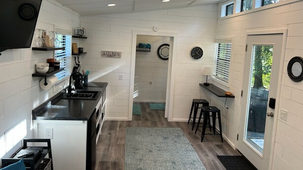 A modern kitchen with white walls, black countertops, and a wooden floor. There are built-in shelves, a clock, and a door with a window. - Tiny House for two nestled under massive oaks in Southern Oklahoma. (Lone Grove)
