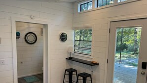 A clean, modern kitchen with white walls, a wooden floor, a window with blinds, two black stools, and a small countertop. - Tiny House for two nestled under massive oaks in Southern Oklahoma. (Lone Grove)