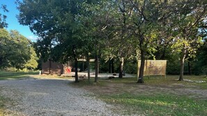 A gravel parking area with trees and a fenced area in the background. - Tiny House for two nestled under massive oaks in Southern Oklahoma. (Lone Grove)